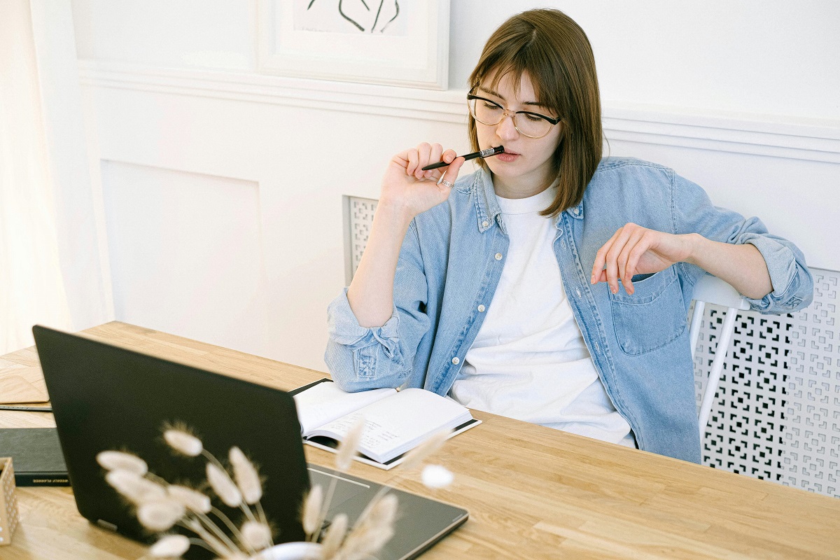 writer sitting at a desk with a notebook and a laptop, holding a pen thoughtfully against her lips